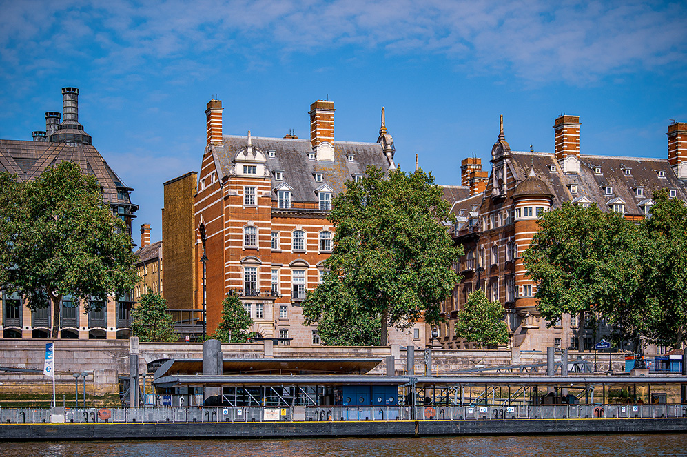 The image depicts a row of historic buildings along a riverbank, featuring classic architectural elements such as chimneys, dormer windows, and decorative facades. The buildings are surrounded by lush green trees and a modern structure, possibly a pier or platform, is visible in the foreground along the river's edge.
