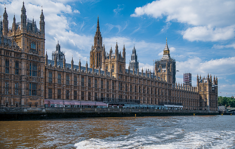 The image depicts the Palace of Westminster, located in London, UK. This iconic building houses the Houses of Parliament and is situated on the north bank of the River Thames. The architecture is characterized by its intricate Gothic design, featuring pointed spires, ornate detailing, and large windows. The sky is partly cloudy, and the river is visible in the foreground, adding to the scenic view.