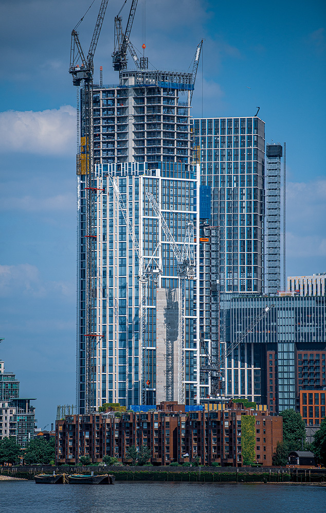 The image depicts a tall building under construction in an urban setting. The structure is surrounded by cranes and scaffolding, indicating ongoing development. The building is situated near a body of water, with smaller residential buildings in the foreground. The sky is clear with a few clouds, suggesting a bright day. The overall scene reflects a bustling cityscape with significant architectural development.