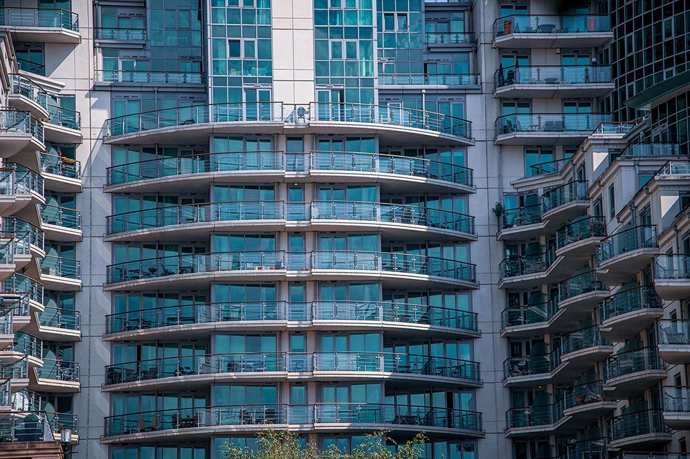 The image depicts a modern, multi-story building with numerous balconies. The balconies are arranged in a curved, wave-like pattern, and each one is enclosed with glass railings. The building's facade features large, blue-tinted glass windows. The design is contemporary, with clean lines and a repetitive pattern that gives it a rhythmic appearance. The structure appears to be a residential or mixed-use building, likely an apartment complex or condominium.