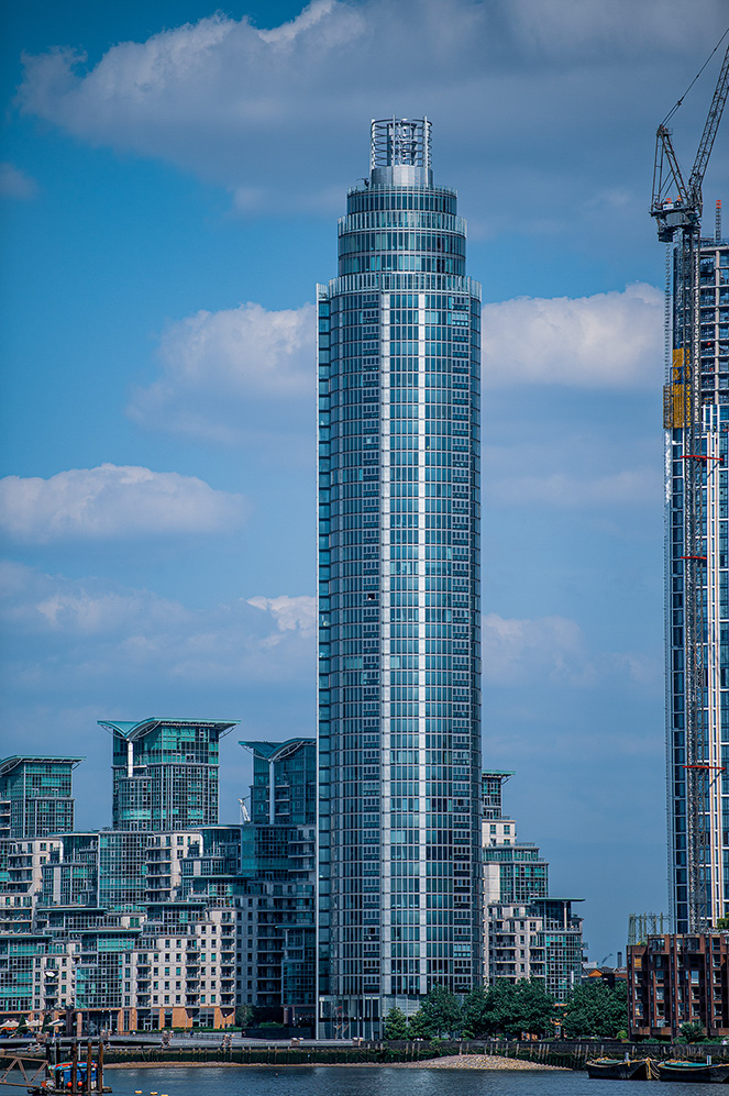 The image showcases a modern urban landscape featuring a tall, cylindrical skyscraper as the central focus. This skyscraper is surrounded by various other buildings, including mid-rise structures with a mix of glass and concrete facades. The scene is set against a backdrop of a partly cloudy sky, with the tall building reflecting the sunlight, giving it a bright and prominent appearance. Additionally, there is a construction site visible on the right side of the image, indicating ongoing development in the area. The foreground includes a body of water, likely a river or canal, with some greenery and a few smaller structures along the waterfront.