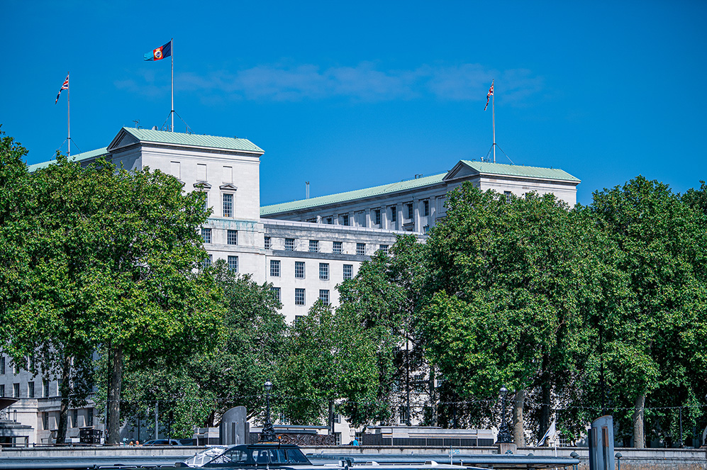 The image depicts a large, white building with a green roof and multiple flags flying atop flagpoles. The building is surrounded by lush, green trees and is situated along a river, with cars and a boat visible in the foreground.