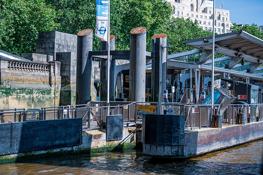 The image depicts a ferry terminal with a ramp for vehicle and pedestrian access. The structure includes metal railings, columns, and a covered waiting area. There are signs indicating the terminal's purpose and directions. The terminal is situated next to a water body, with greenery and buildings visible in the background.