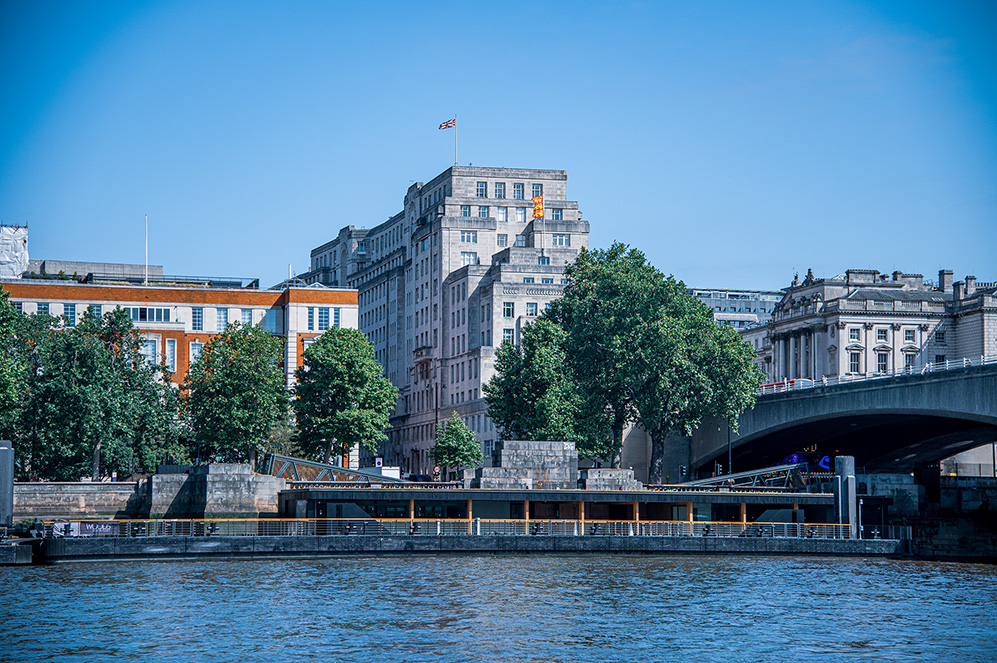 The image depicts a cityscape with a river in the foreground. Prominent buildings, including a tall structure with a flag on top, line the riverbank. There are several trees along the river, and a bridge extends across the water. The sky is clear, and the overall scene suggests a calm, urban environment.