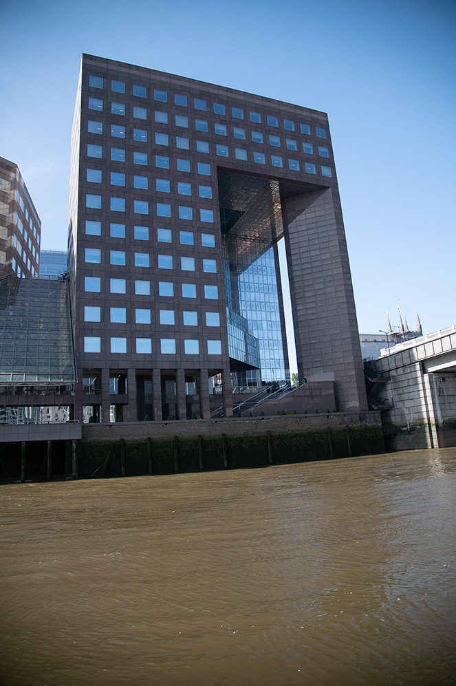 The image depicts a modern architectural building with a distinctive design, featuring a large rectangular structure with a grid of windows and a prominent vertical opening in the center. The building is situated next to a body of water, with a clear blue sky in the background. The design includes a mix of glass and concrete materials, and the building is surrounded by other modern structures.