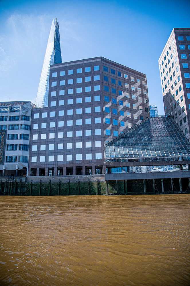 The image depicts modern buildings, including a distinctive glass skyscraper and a building with a grid-like facade, situated near a body of water. The architecture is contemporary, with a mix of glass and metal materials, and the scene is set on a clear, sunny day.