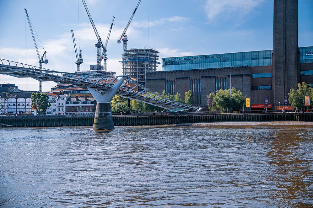 The image depicts a riverside scene with a modern pedestrian bridge extending from the shore into the water. In the background, there is a large building with a sign that reads 'SEE GREAT ART FROM AROUND THE WORLD,' indicating it is likely a museum or gallery. Several construction cranes and a building under construction are visible, suggesting ongoing development in the area. The scene includes a mix of modern and older architecture, with trees and a clear sky overhead.