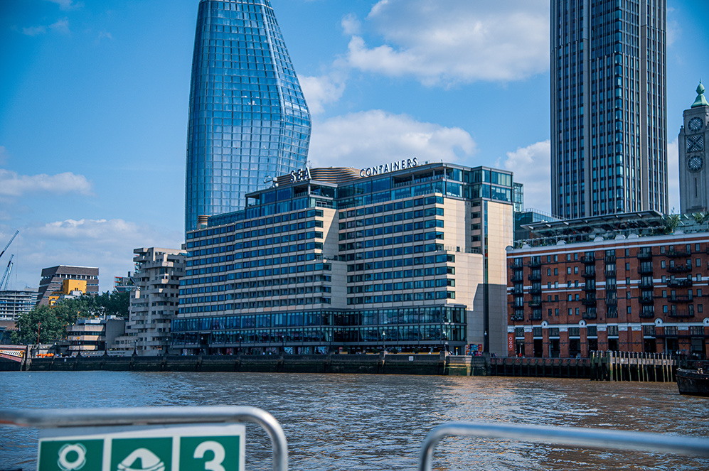 The image depicts a modern urban waterfront scene featuring contemporary architecture. Prominent buildings include a distinctive, curved glass skyscraper and a mid-rise building labeled 'CONTAINERS'. The setting appears to be a bustling city with a mix of older brick buildings and newer glass structures. The waterfront area includes a dock with a sign indicating a life preserver and the number 3, suggesting it is a designated safety or docking area. The sky is clear with some clouds, indicating a bright and sunny day.