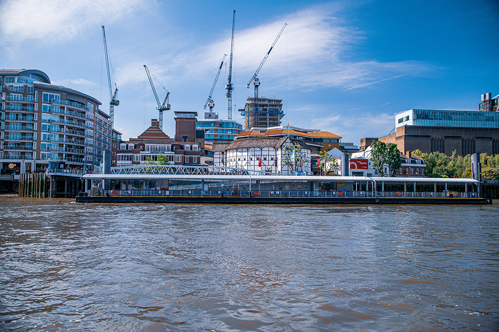 The image depicts a riverside urban scene with a mix of modern and historic architecture. There are several construction cranes in the background, indicating ongoing development. The river in the foreground is calm, reflecting the buildings and sky. Notable structures include a large, ornate building with a glass and metal facade, and a modern building with the words 'Great Art' visible on its side. The scene is set under a clear blue sky with some wispy clouds.