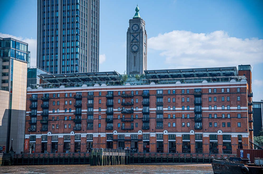 The image depicts a waterfront scene with a prominent red-brick building in the foreground. This building has multiple floors with balconies and large windows. Behind it, there are modern high-rise buildings, including a tall clock tower with a green spire. The sky is clear with a few scattered clouds. The overall setting suggests an urban environment, possibly a city with a mix of historical and contemporary architecture.