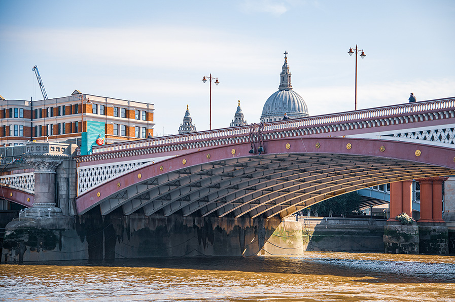 TThe image depicts a bridge with ornate architectural details, spanning a body of water. In the background, there is a large domed building with smaller domes, likely a significant landmark. The bridge features decorative elements and is supported by large stone pillars. There are buildings on the left side, and the scene is set under a clear sky with some clouds.