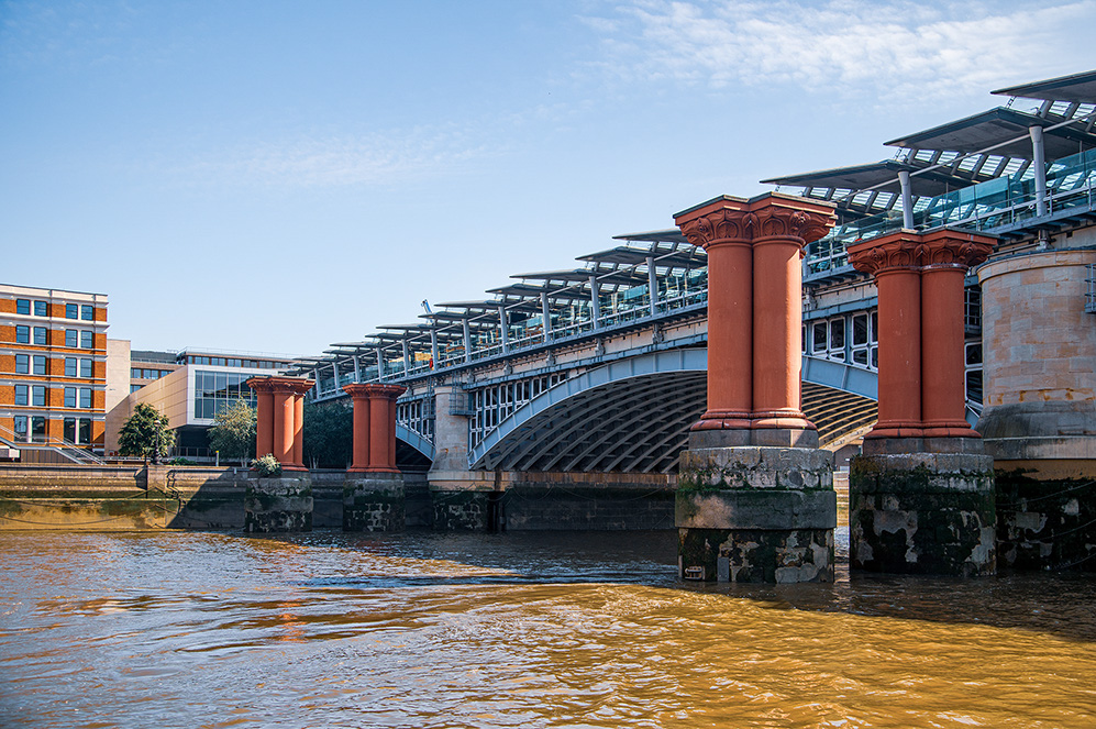 The image shows a bridge with distinctive architectural features, including large, classical-style columns supporting its structure. The bridge spans over a body of water and has a modern, industrial design with a metal framework and solar panels on top. The surrounding area includes modern buildings and a clear blue sky, suggesting a well-developed urban environment.