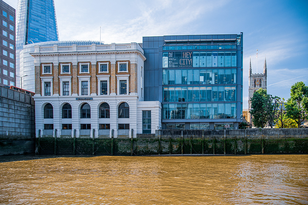 The image depicts a riverside view of modern and historic buildings in an urban setting. The scene includes a mix of architectural styles, with a prominent glass-fronted building displaying a 'HEY CITY' sign. Adjacent to it is a more traditional building with arched windows and a brick facade. The river in the foreground adds a natural element to the urban landscape, and a church spire is visible in the background, indicating the presence of historical architecture in the vicinity.
