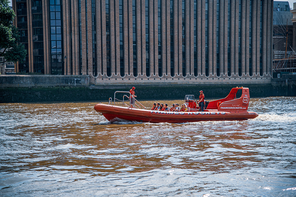 The image shows a red speedboat named 'Thames Rockets' on a river, with several passengers and two crew members on board. The boat is moving away from the viewer, and the background features a large, modern building with vertical windows and a concrete embankment.