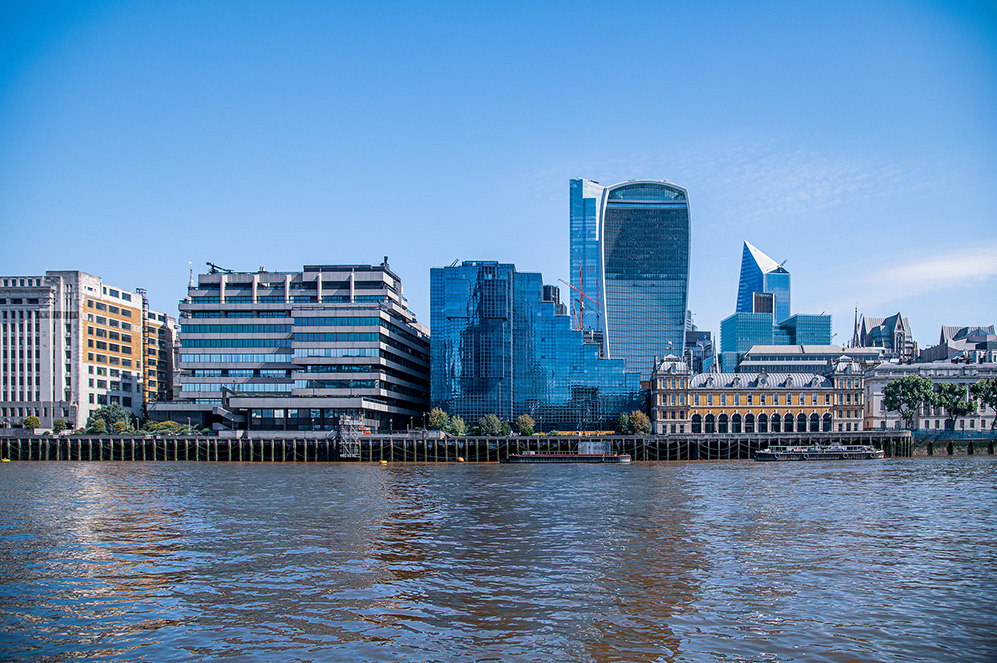 The image depicts a modern cityscape featuring a mix of contemporary skyscrapers and historic buildings along a riverbank. Notable structures include the Walkie-Talkie building and other distinctive high-rises, showcasing a blend of architectural styles. The river in the foreground adds a serene element to the bustling urban scene.