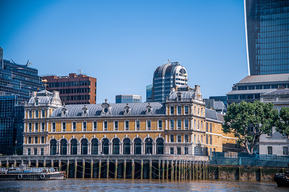 The image depicts a historic building with a distinctive architectural style, featuring arched windows and a domed roof, situated along a river. The building is surrounded by modern skyscrapers, indicating an urban setting. The contrast between the old and new architecture is notable. A boat is docked nearby, and the scene is set under a clear blue sky.