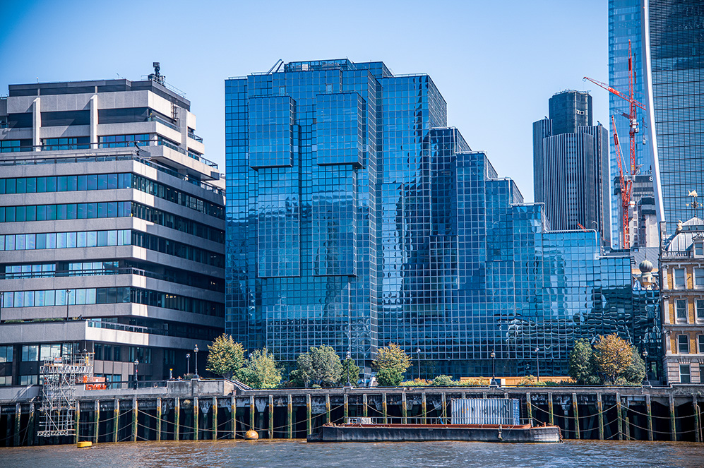 The image depicts a modern cityscape featuring a mix of contemporary skyscrapers and older architecture. The skyscrapers are primarily made of glass, reflecting the blue sky and surrounding buildings. There is a river or water body in the foreground, with a dock and some greenery along the waterfront. The scene suggests a bustling urban environment with ongoing construction, as indicated by the presence of a crane.