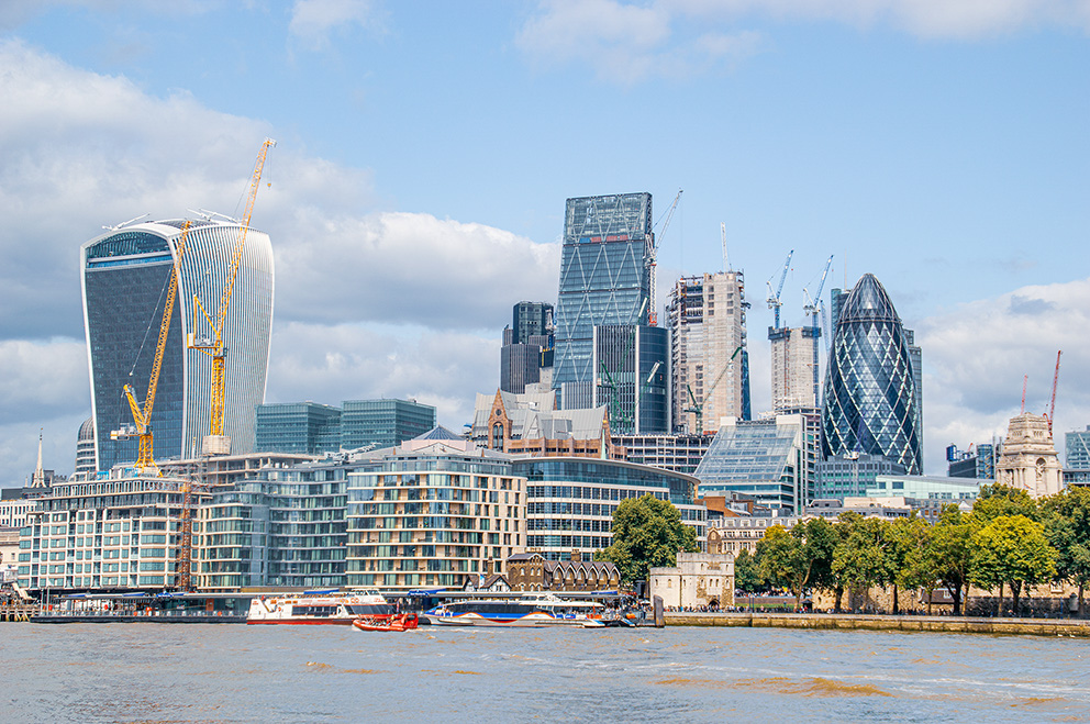The image depicts a modern cityscape with a river in the foreground. Prominent features include several distinctive skyscrapers, such as the Walkie-Talkie and the Gherkin buildings. There are also construction cranes indicating ongoing development. The scene is lively with boats on the river and a mix of architectural styles, blending contemporary and historical elements.
