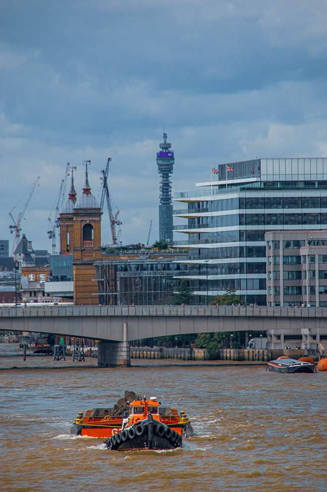 The image depicts a bustling urban scene with a river in the foreground. A barge is seen on the river, loaded with construction materials. In the background, there is a mix of modern and historic architecture, including a large bridge, cranes, and a distinctive tower. The sky is overcast, suggesting a cloudy day. The overall setting appears to be a city undergoing development, likely a major metropolitan area.