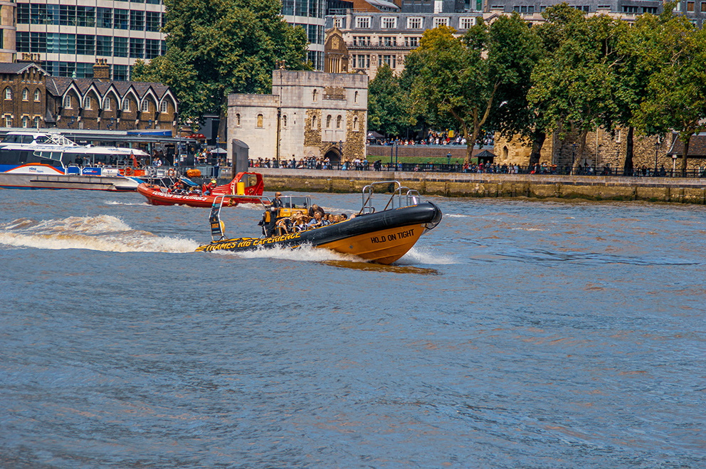 The image depicts a scene on a river with several boats, including a prominent black and yellow inflatable boat labeled 'HOLD ON TIGHT' with 'THAMES RIB EXPERIENCE' written on it. The boat is moving quickly, creating a wake behind it. In the background, there is a historic stone building, likely a castle or fortress, with people gathered around it. The surrounding area includes modern buildings and lush green trees, indicating an urban setting with historical significance.