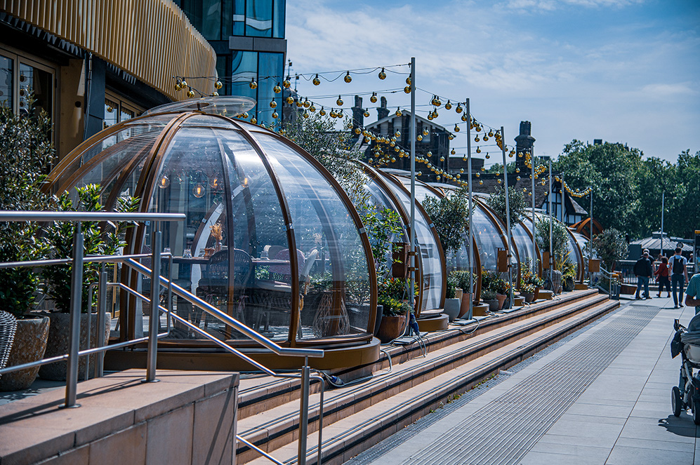 The image depicts a modern outdoor dining setup with transparent, dome-shaped structures along a waterfront. Each dome houses a dining table and chairs, providing a unique and intimate dining experience. The area is adorned with string lights, adding a festive and cozy atmosphere. The setting includes potted plants and a railing along the waterfront, with people walking and enjoying the surroundings. The overall ambiance suggests a blend of contemporary design and natural beauty.
