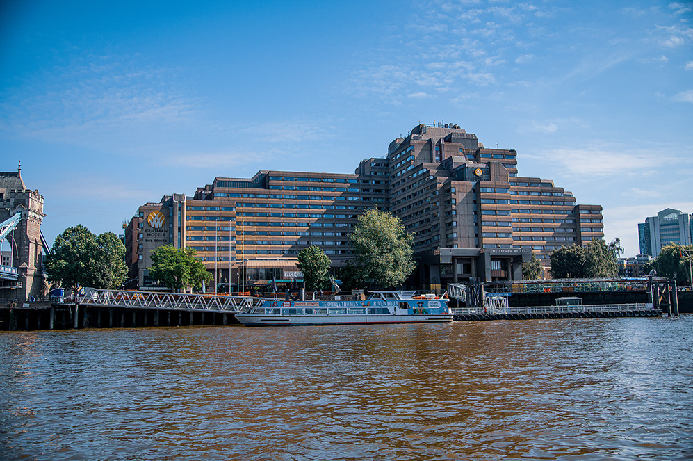 The image depicts a large, multi-story hotel named 'The Tower Hotel' situated along a river. The hotel has a distinctive, modern architectural design with multiple wings and balconies. In front of the hotel, there is a dock with a boat named 'Buquebus' moored, indicating the availability of water transportation services. The scene is set on a clear day with a blue sky and some clouds. The surrounding area includes trees and other buildings, suggesting an urban setting.