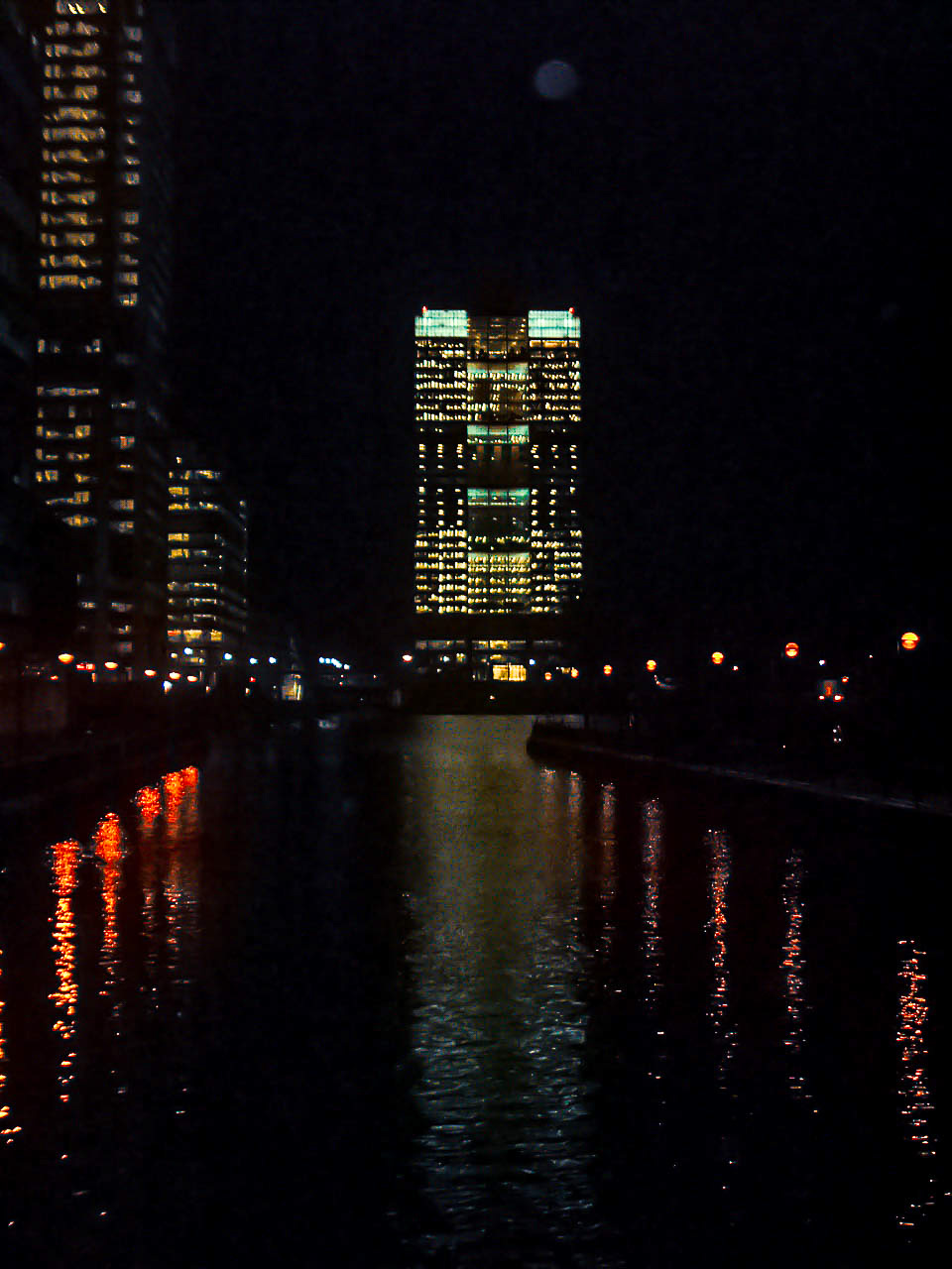 The image depicts a nighttime cityscape with tall buildings illuminated by lights. The reflections of the lights can be seen in the water in the foreground, creating a visually striking scene. The sky is dark, and the moon is visible, adding to the overall ambiance of the photograph.