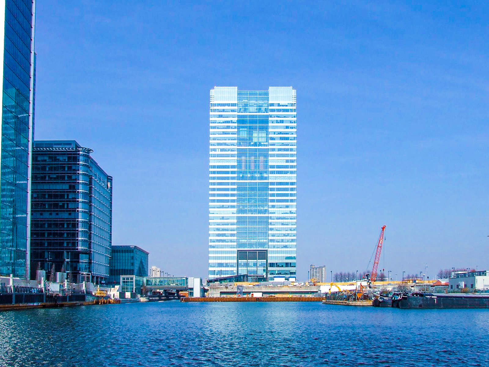 The image depicts a waterfront scene with modern buildings and a construction site. The tallest building in the center has a reflective glass facade, and there are other high-rise buildings nearby. The water in the foreground suggests that this area is likely a harbor or a riverfront. The construction site includes cranes and various construction materials, indicating ongoing development.