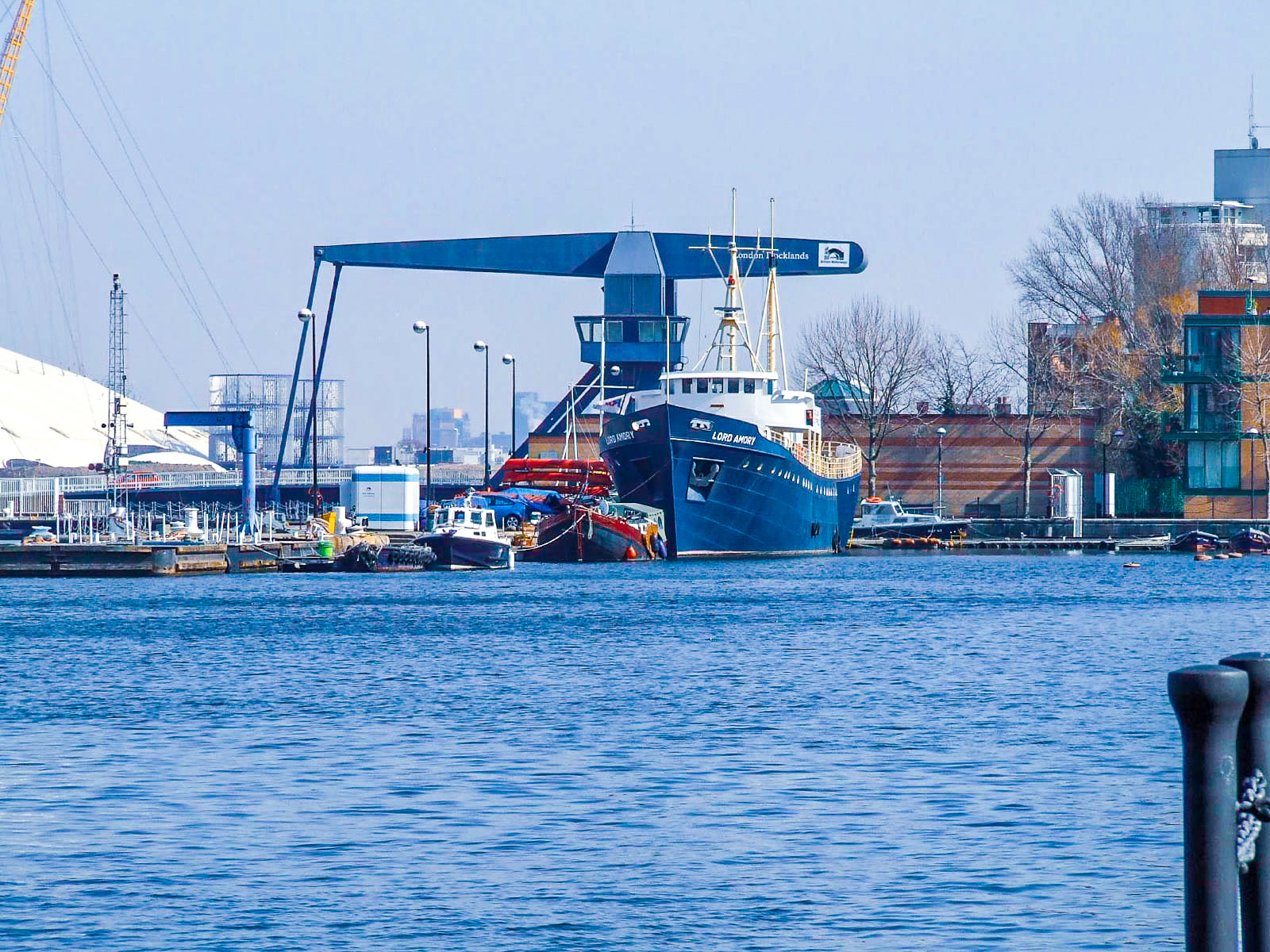 The image shows a large ship named 'Lord Ashfield' docked at a port. The ship is blue and white and has 'London Docklands' written on it. There are smaller boats and various structures around the ship, and the scene appears to be in an industrial or commercial dock area with buildings and cranes in the background.