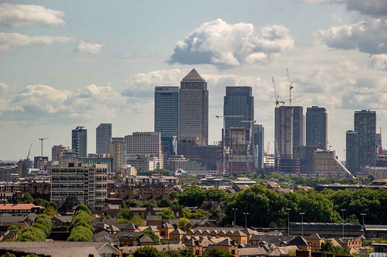 The image depicts a cityscape featuring a mix of high-rise buildings and residential areas. Prominent skyscrapers, including those with the logos of HSBC and Citi, dominate the skyline. Construction cranes and ongoing construction projects indicate significant development activity. The foreground shows a contrast between the modern skyscrapers and the older, smaller residential houses and buildings. The sky is partly cloudy, adding to the overall urban atmosphere.