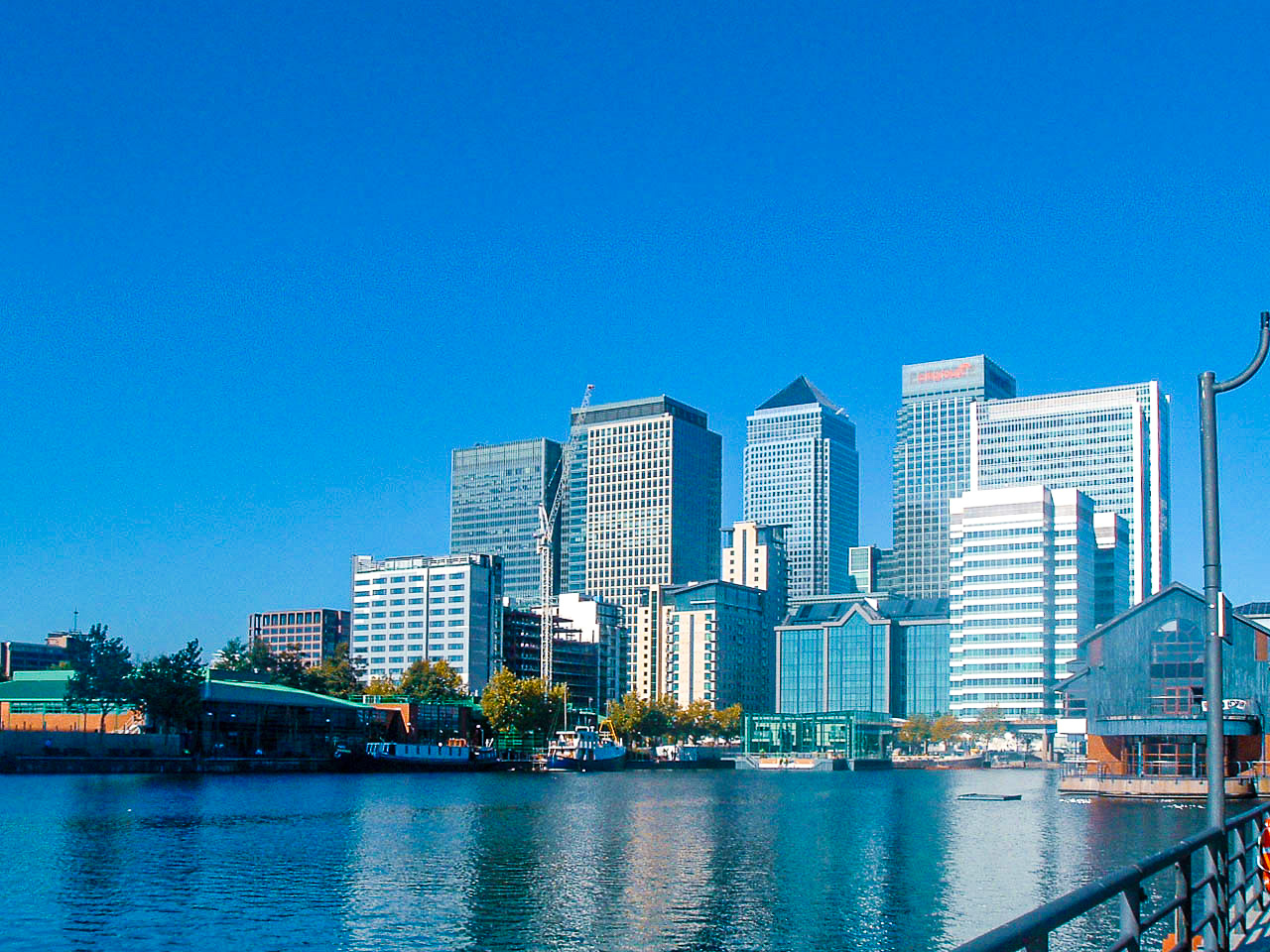 The image depicts a modern cityscape with tall skyscrapers and office buildings along a waterfront. The buildings are reflective and contemporary in design, with a mix of glass and steel structures. The sky is clear and blue, indicating a sunny day. In the foreground, there is a body of water with some boats and a dock area. The scene suggests a bustling urban environment, likely a financial or business district.