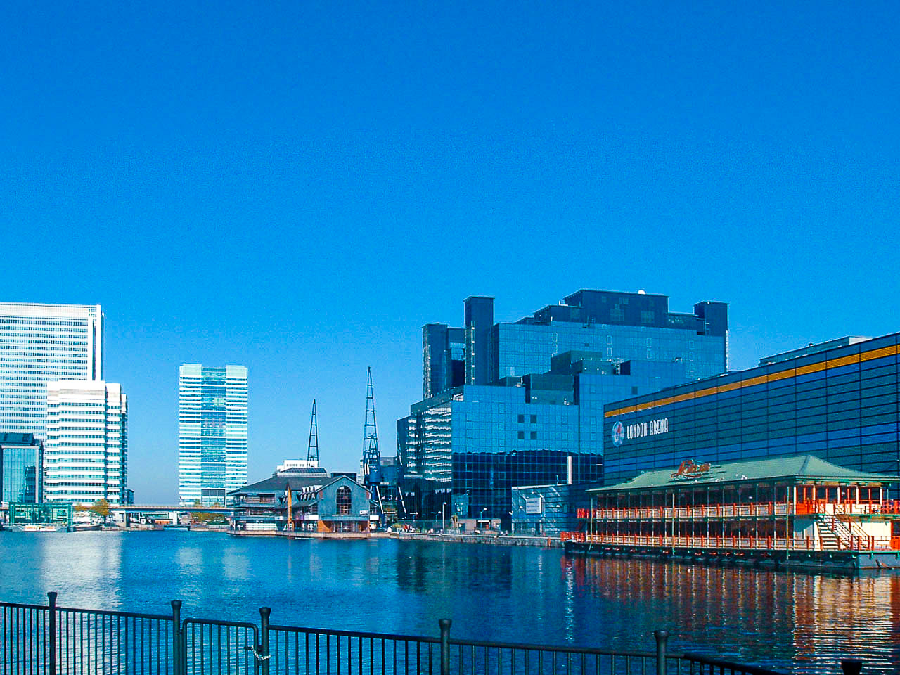 The image depicts a waterfront urban scene with modern buildings and a clear blue sky. The London Arena is prominently visible on the right side of the image. The scene includes a mix of high-rise office buildings and a historic-looking structure near the water. The water reflects the buildings, adding to the scenic view. The area appears to be a blend of commercial and recreational spaces.