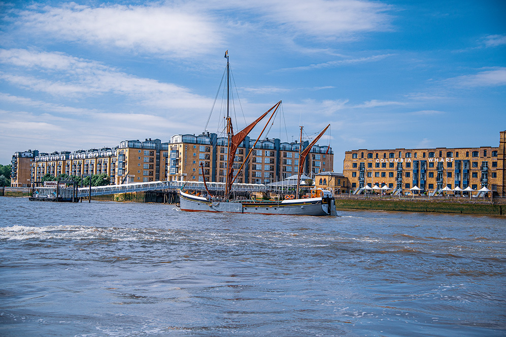 The image shows a scenic view of a waterfront area with a traditional sailing boat docked near a modern building complex. The buildings are multi-story with a mix of brick and glass architecture, and there is a sign that reads 'OMAHA WHARF.' The sky is clear with some clouds, and the water appears calm, reflecting the structures and the boat.