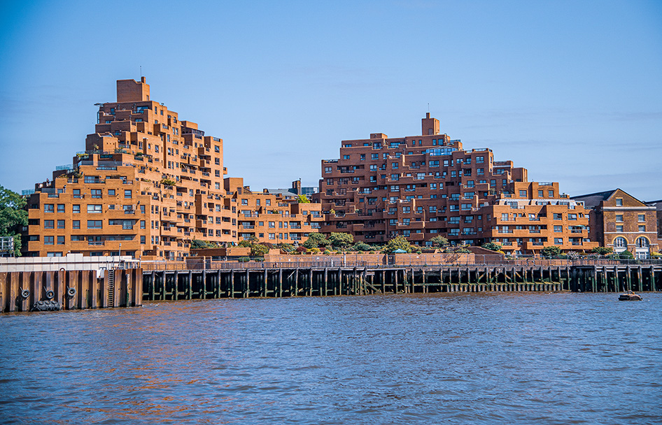 The image depicts a waterfront view of a distinctive residential complex with a unique architectural design, characterized by a series of stacked, terraced buildings with a brick facade. The buildings are situated along a water body, with a wooden pier extending into the water in the foreground. The sky is clear, and the overall scene suggests a calm, urban waterfront environment.