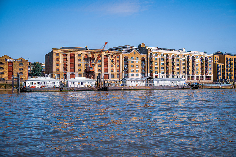 The image depicts a waterfront scene with historic brick buildings along the shore. The buildings have a mix of architectural styles, including large arched windows and a combination of brick and stone facades. There are several boats docked along the water's edge, and a crane is visible, suggesting ongoing construction or maintenance. The water is calm, reflecting the buildings and the clear blue sky above. The overall atmosphere is serene and picturesque, typical of a revitalized waterfront area.