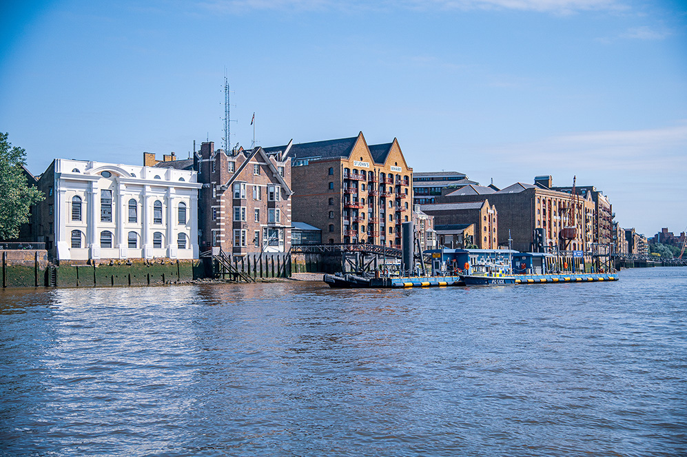 The image depicts a waterfront area with historic buildings and a river. The buildings are a mix of architectural styles, including a prominent white building with large windows and several brick buildings with gabled roofs. There is a small dock with a boat moored alongside, and the scene is bathed in daylight under a clear blue sky.