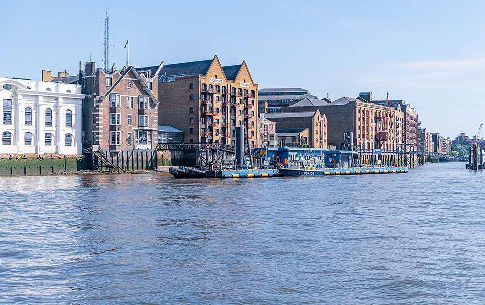 The image depicts a waterfront scene with historic buildings and a docked boat. The buildings are a mix of architectural styles, featuring brick facades and large windows. The boat is moored at a wooden pier, and the water is calm, reflecting the structures along the shore. The sky is clear, indicating a sunny day. The overall atmosphere is serene and picturesque, typical of a harbor or riverside area.