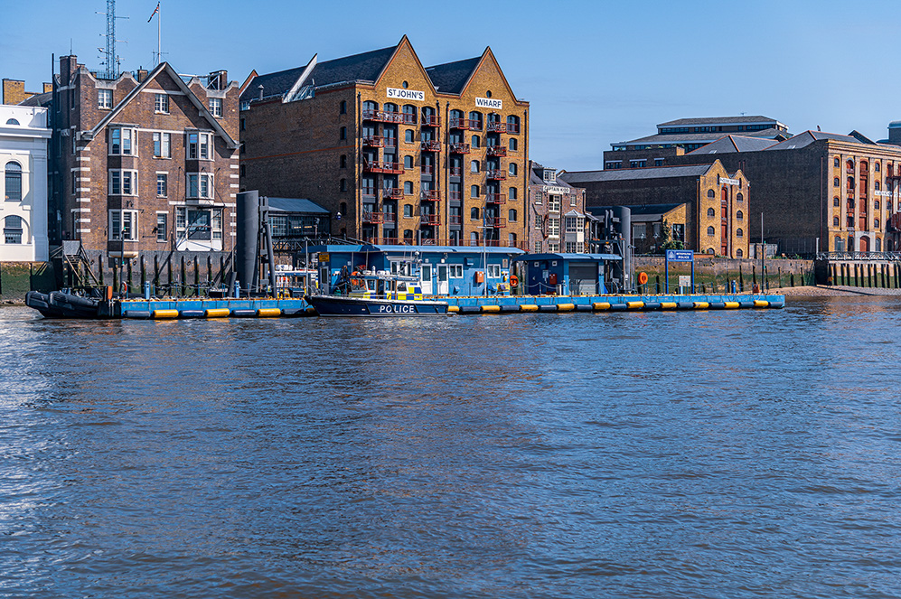The image depicts a waterfront scene with historic brick buildings, including one labeled 'St. John's Wharf.' In the foreground, there is a police boat docked at a floating pier. The area appears to be a mix of residential and commercial buildings, with a clear sky and calm water.