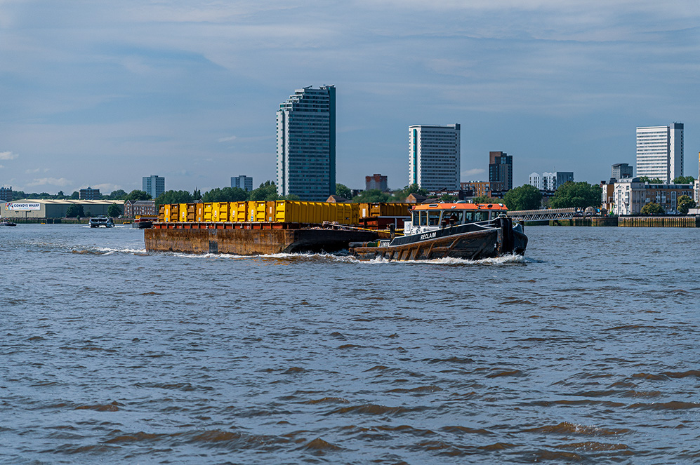 The image shows a barge loaded with yellow containers being towed by a tugboat on a river. The scene is set against a backdrop of modern high-rise buildings and other structures, indicating an urban environment. The water is slightly choppy, and the sky is partly cloudy, suggesting a typical day with moderate weather conditions.