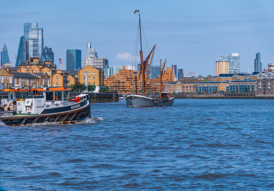 The image depicts a waterfront scene with two boats on a body of water, likely a river. In the foreground, there is a modern tugboat named 'Resource.' In the background, there is a historic sailing ship. The backdrop features a mix of modern skyscrapers and older, more traditional buildings, indicating an urban setting with a blend of architectural styles. The overall atmosphere suggests a bustling, modern city with a rich maritime history.