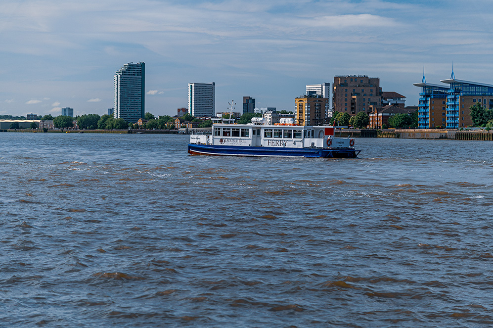 The image shows a river scene with a ferry boat named 'TERRY' in the center, moving through the water. In the background, there is a cityscape with various buildings, including tall modern skyscrapers and shorter, older structures. The sky is partly cloudy, and the overall atmosphere appears calm and serene.