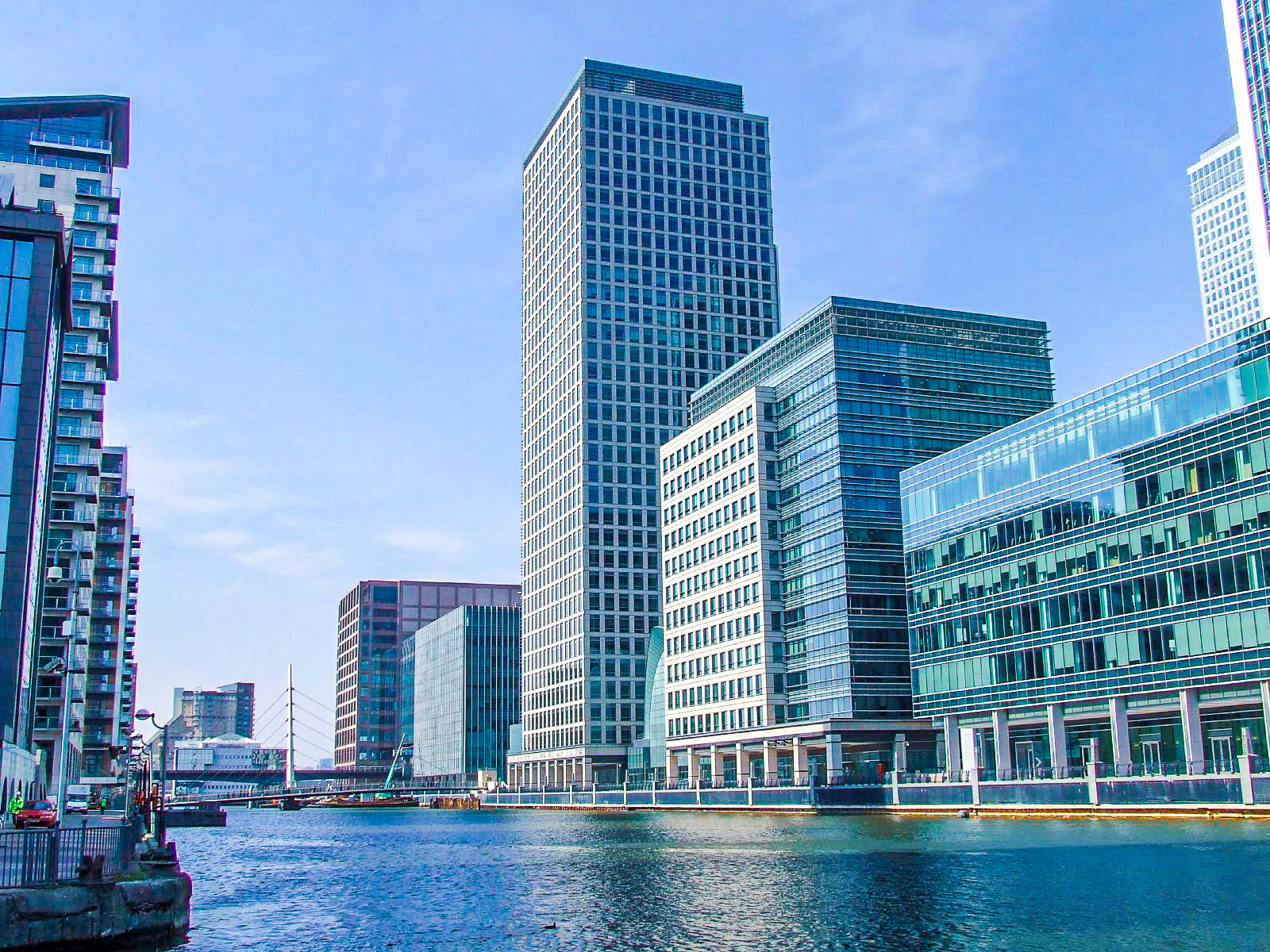 The image depicts a modern waterfront area with several high-rise buildings. The architecture is contemporary, featuring glass and steel structures. The buildings are situated along a body of water, likely a river or canal, with a clear blue sky overhead. The scene suggests a business or financial district, given the corporate appearance of the buildings. The overall atmosphere is bright and vibrant, indicating a clear day.