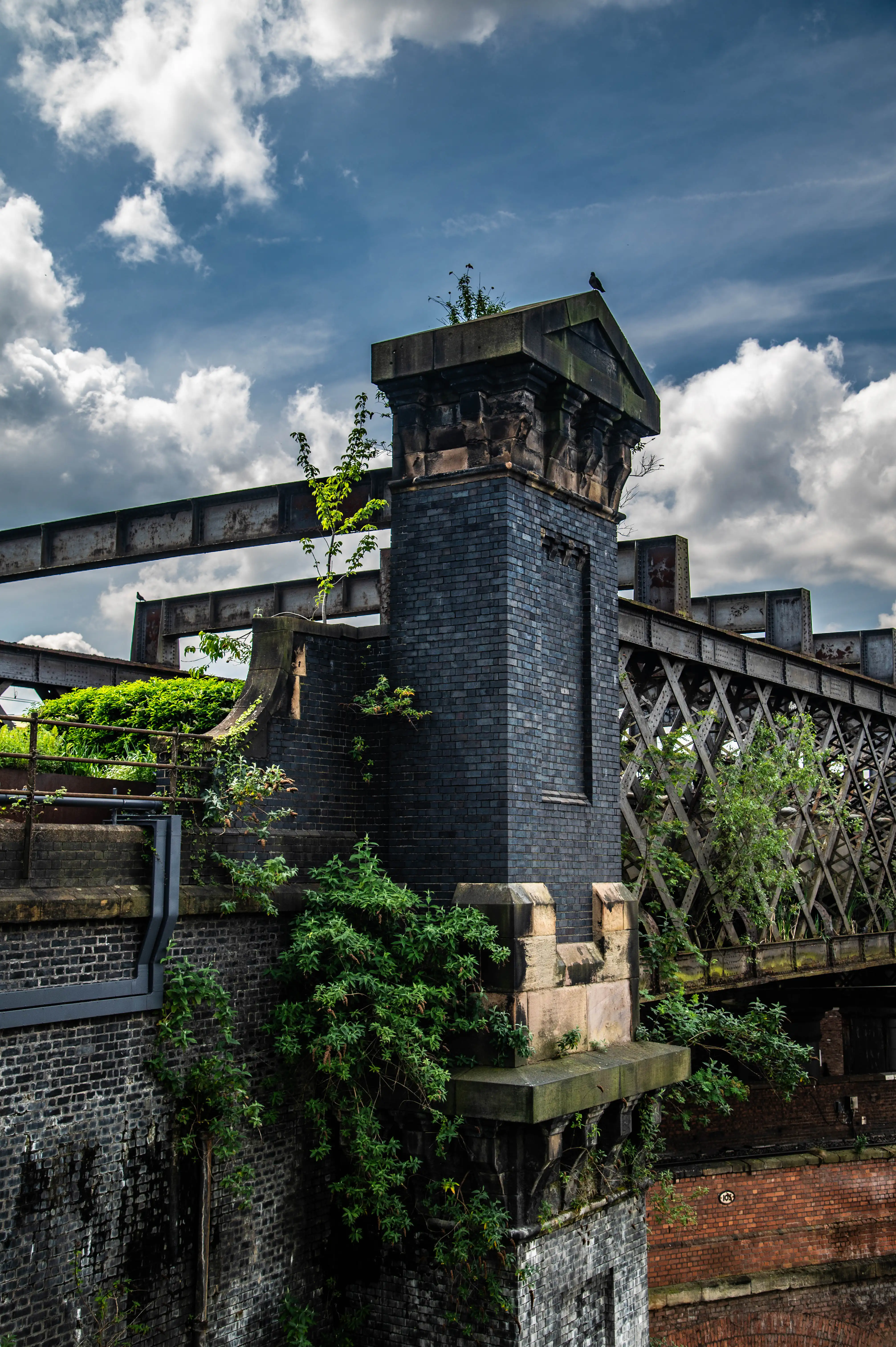 A dark brick pillar topped with ornate stonework stands against a cloudy blue sky. An old, rusted metal railway bridge with latticework is visible behind, intertwined with vegetation.