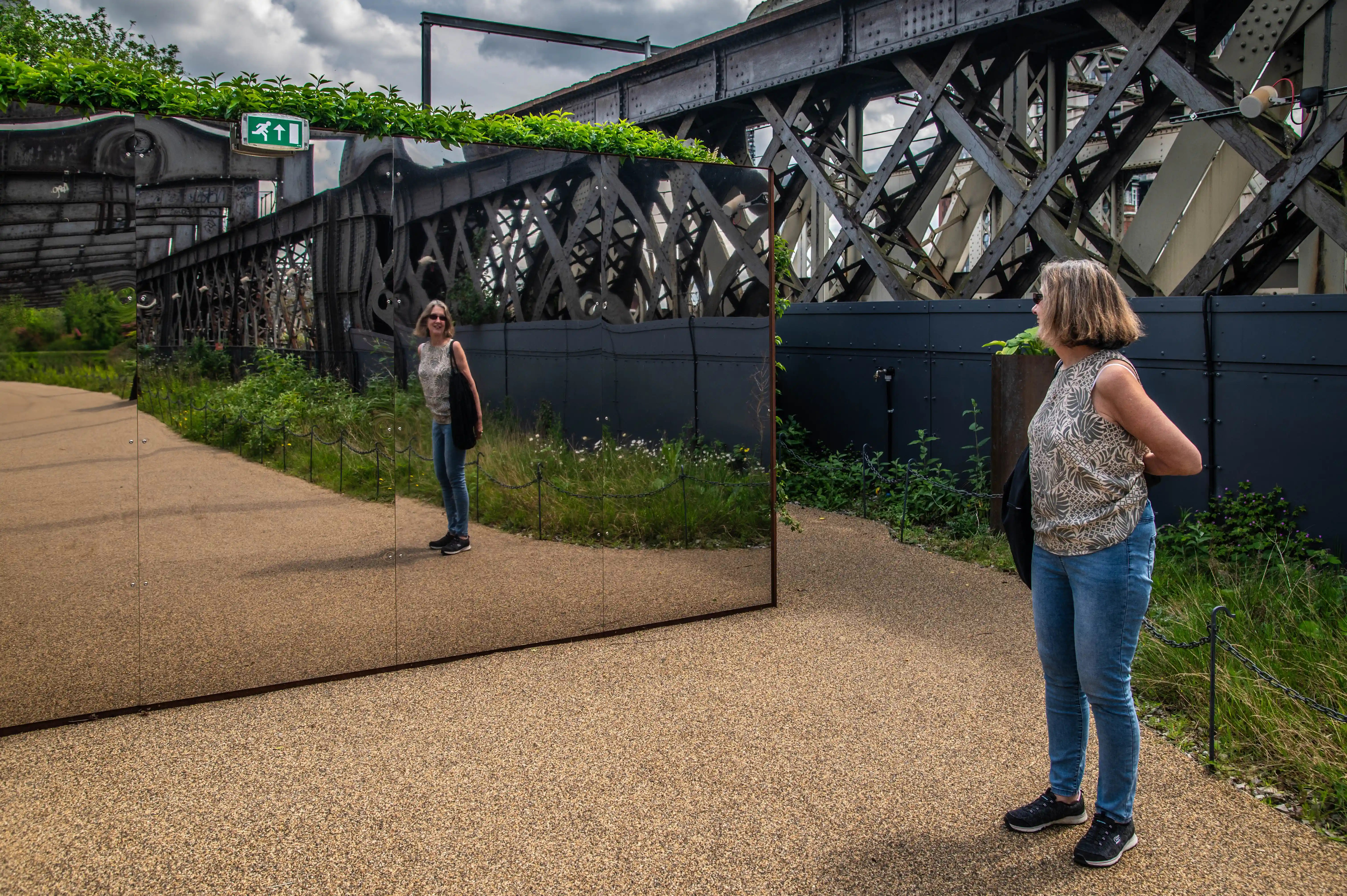 A woman in jeans and a patterned top stands on a gravel path, looking into a large reflective panel. The panel mirrors the intricate metalwork of an overhead bridge structure, lush greenery, and an emergency exit sign.