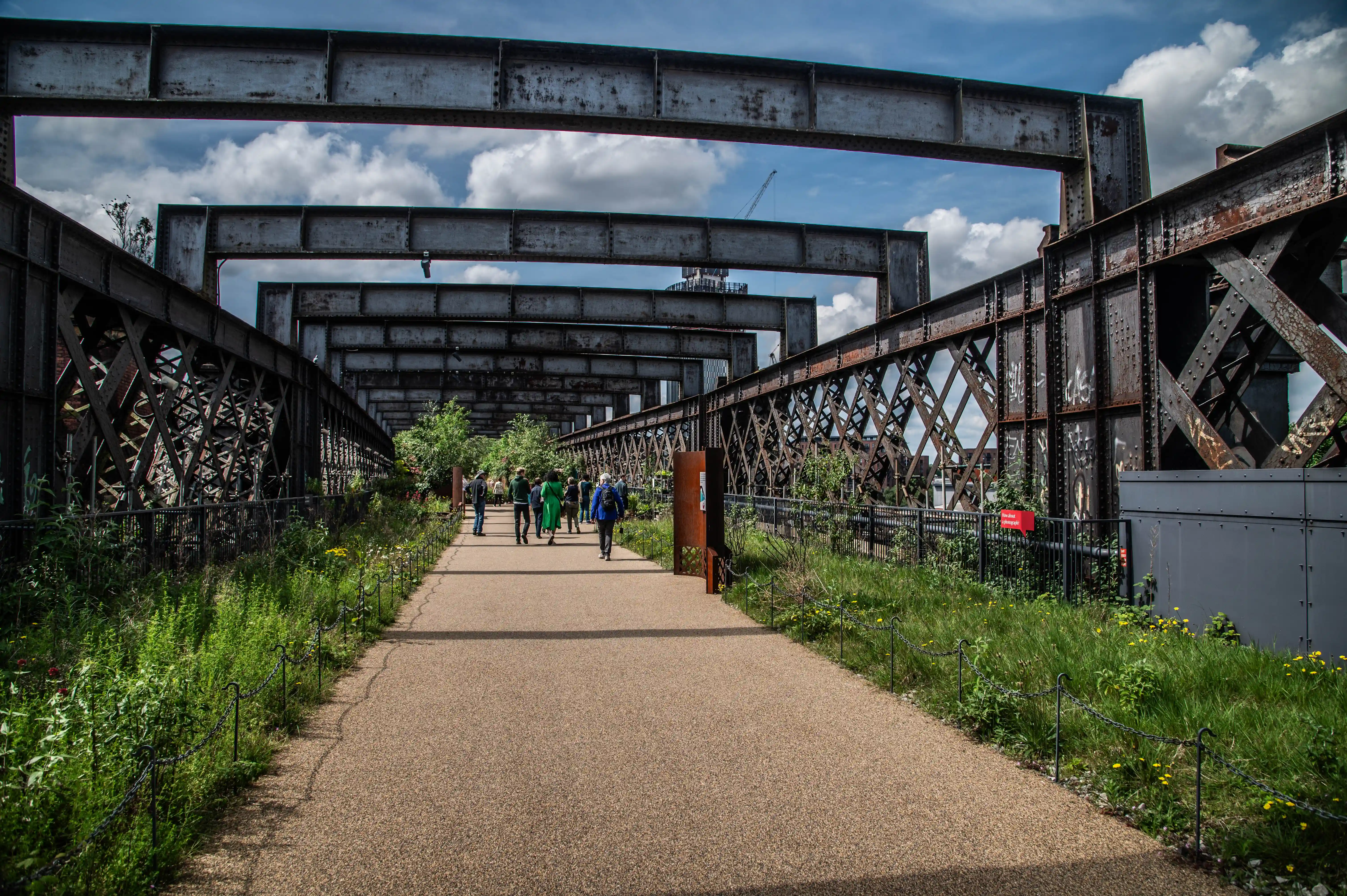 A walkway passes under a series of weathered metal railway bridge sections, with lush vegetation on either side and a few people walking away from the camera.