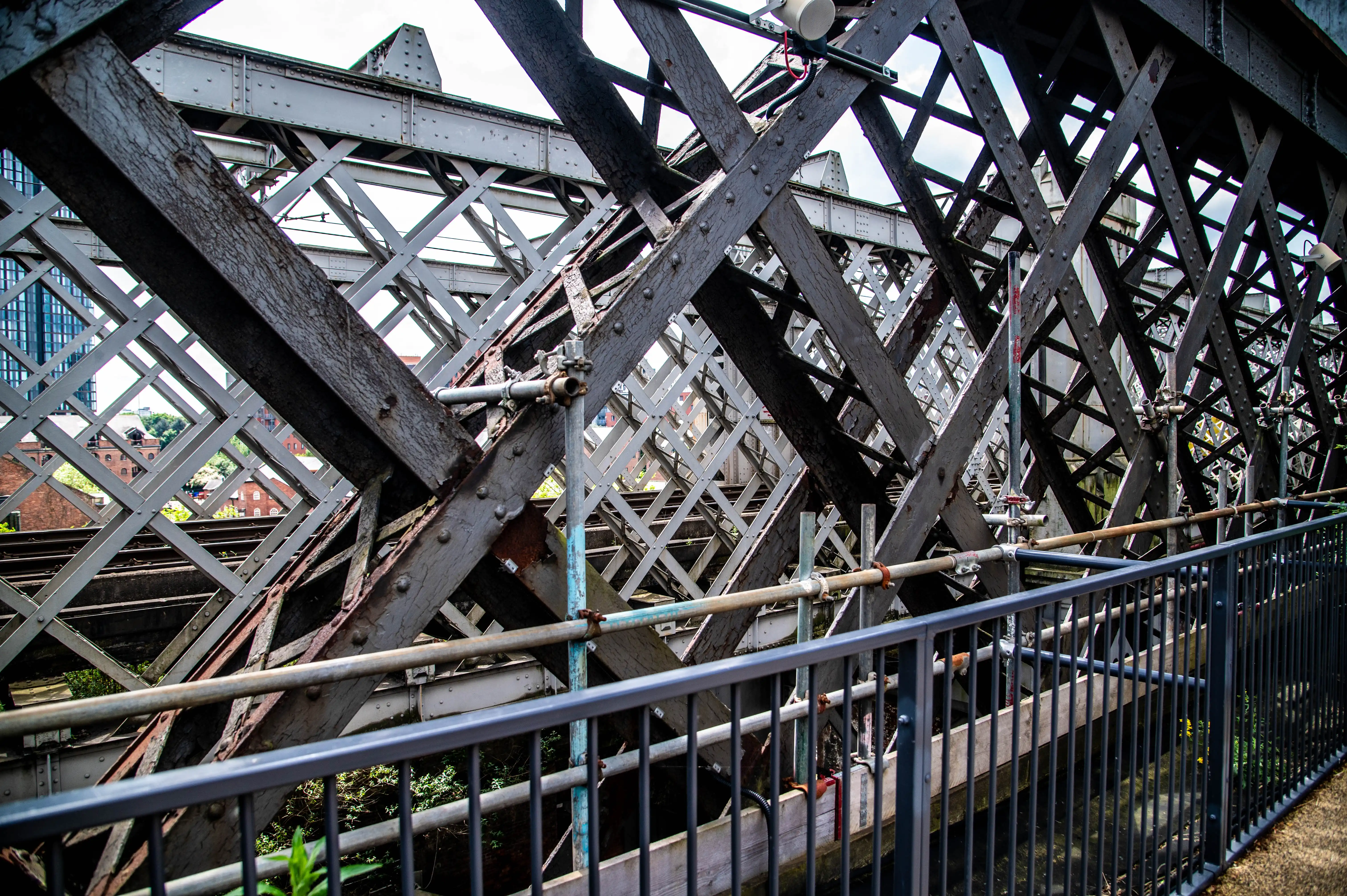 Detailed view of the crisscrossing beams and rivets of an old industrial bridge, with a glimpse of urban buildings and railway tracks in the background.