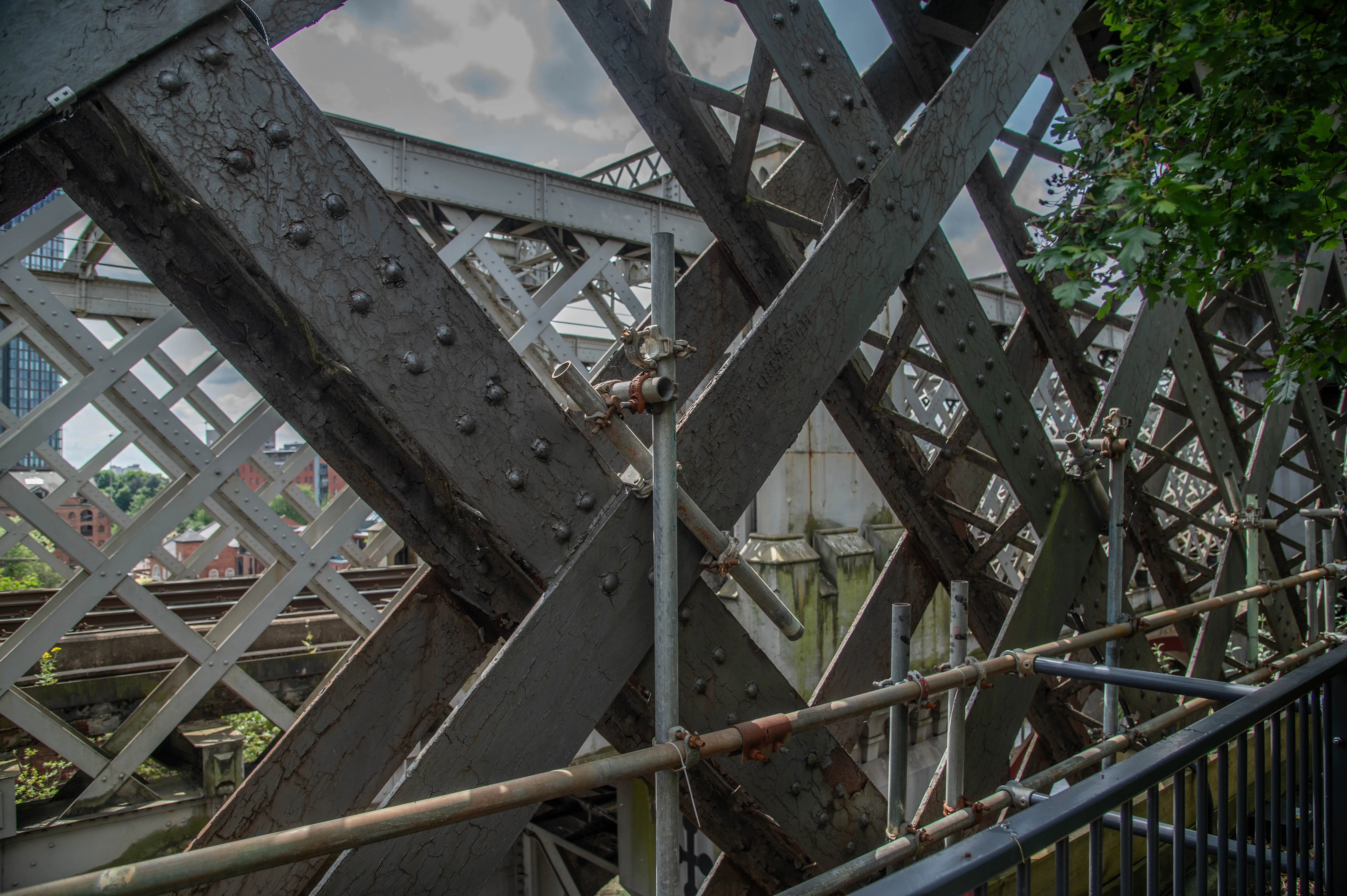 Detailed view of the crisscrossing beams and rivets of an old industrial bridge, with a glimpse of urban buildings and railway tracks in the background.