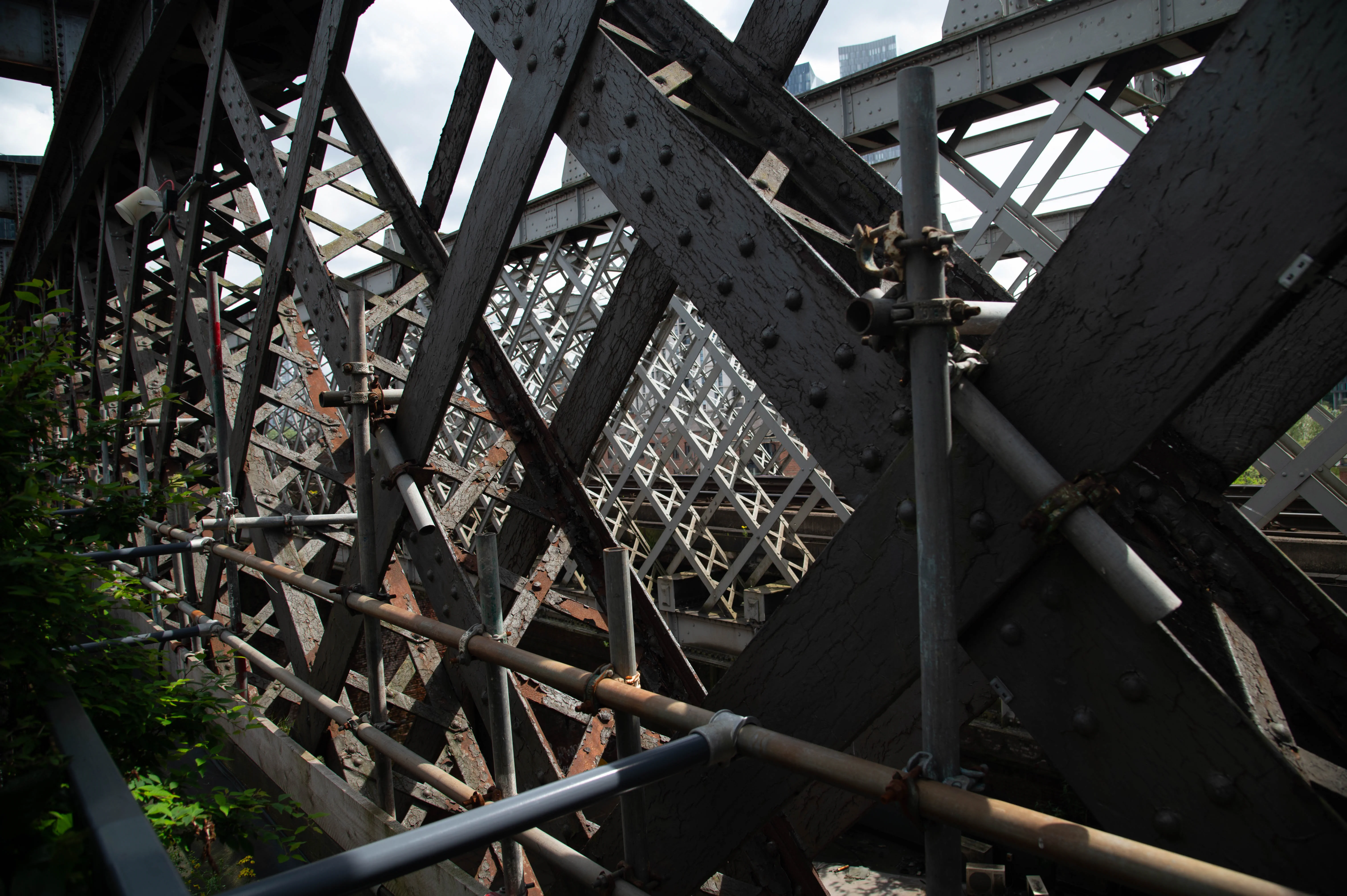 Close-up of a dark grey, riveted iron bridge structure with peeling paint, forming a complex lattice. Scaffolding pipes and clamps are attached to the structure. In the blurred background, railway tracks and brick buildings are visible under a cloudy sky.