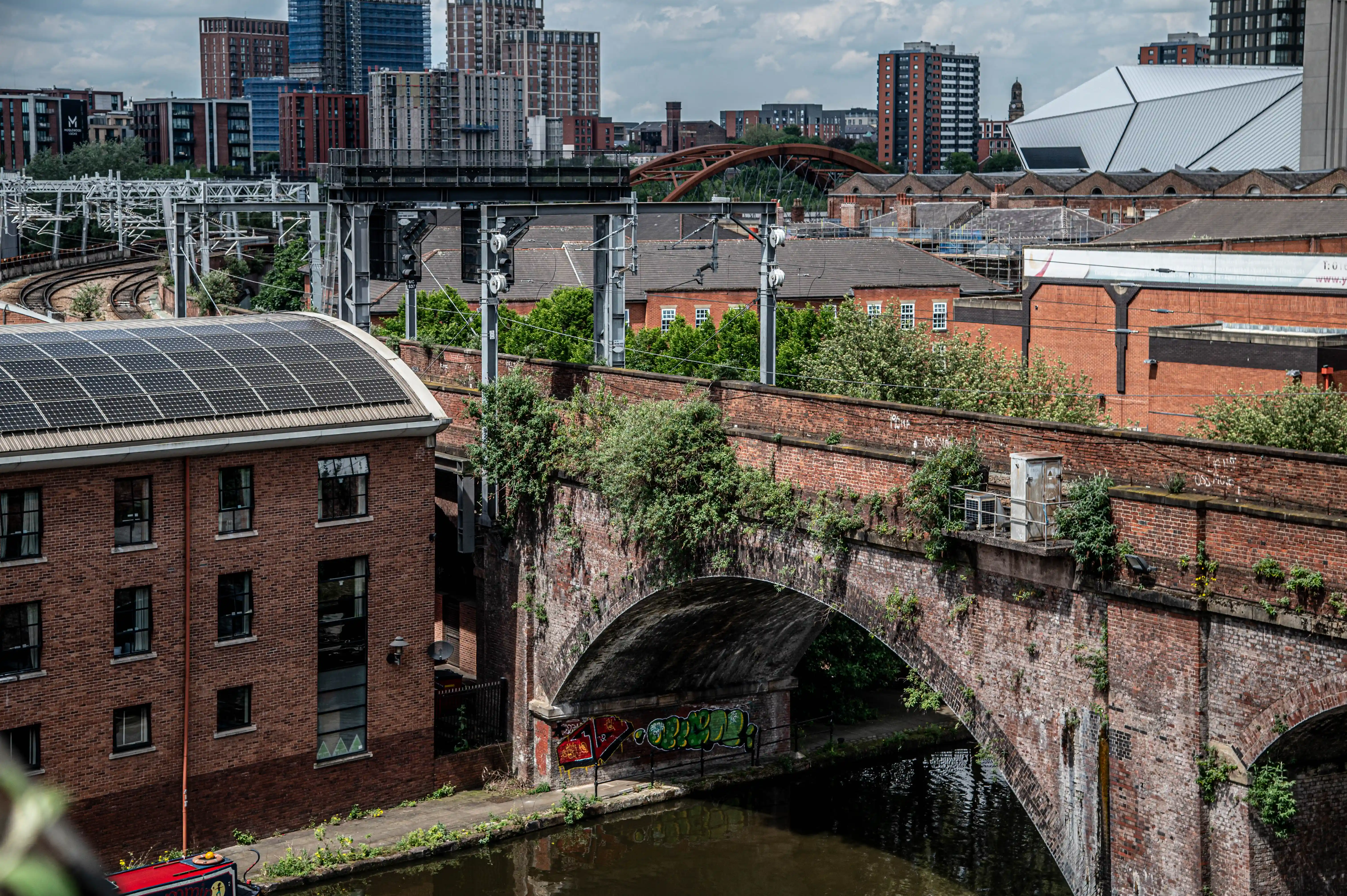 An old brick railway bridge with graffiti arches over a canal, with modern buildings and a stadium in the background.