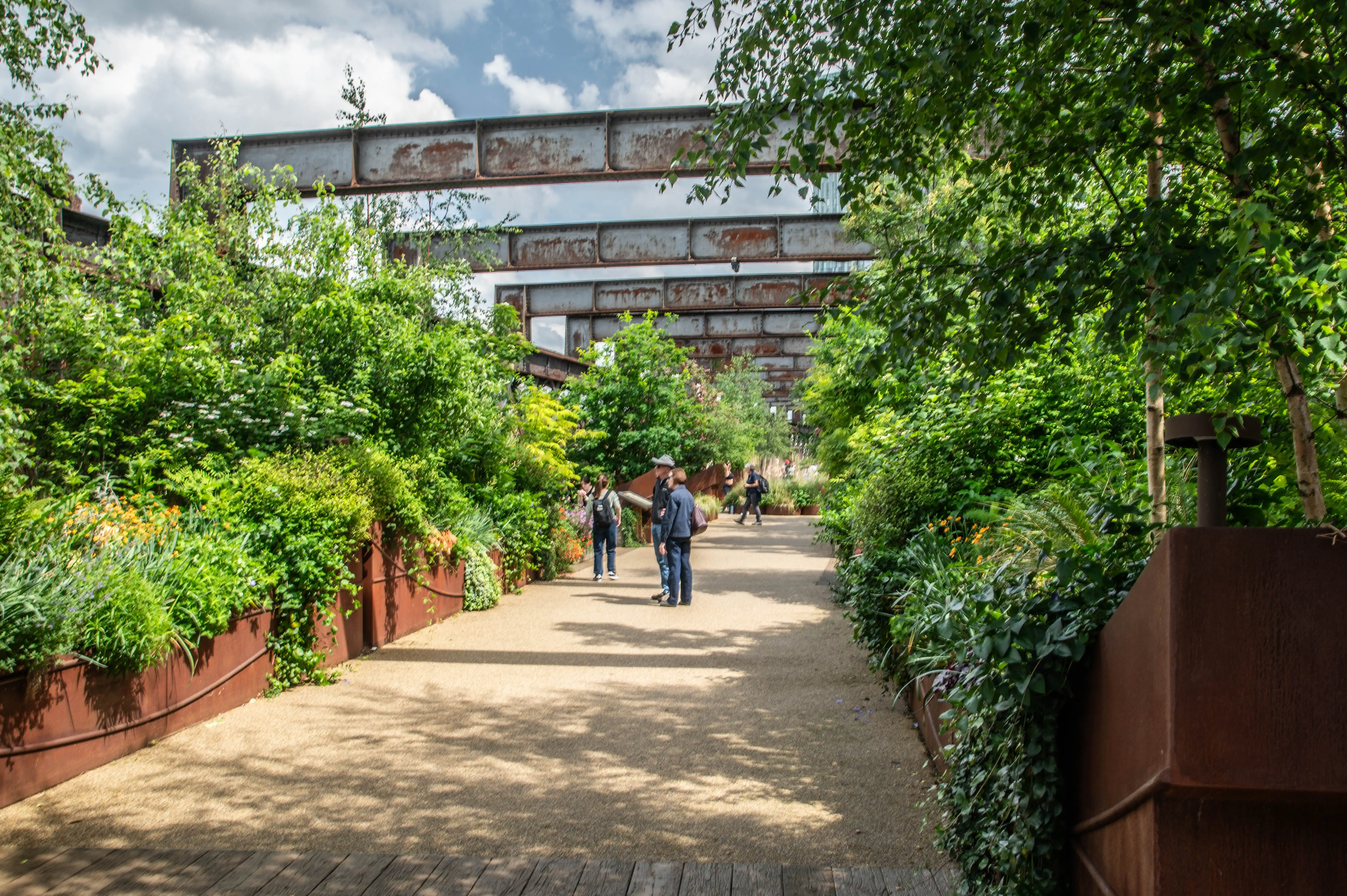 A wide, gravel pathway leads through a green space with lush vegetation on either side. Overhead, large rusty metal beams form an industrial skeletal structure. Several people are walking on the pathway.